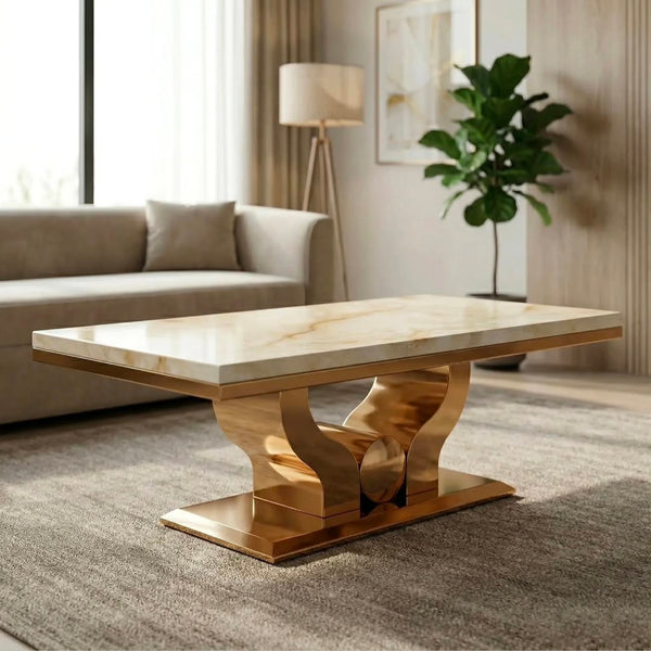 A modern rectangular coffee table with a polished cream marble top and a sculptural metallic gold base, set in a contemporary living room with a sofa, floor lamp, and potted plant.